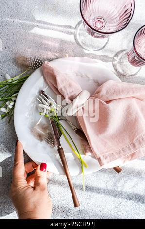 Top view of a cropped unrecognizable woman's hand on an elegant spring table setting featuring snowdrops, a white marble plate, a pink napkin, and vin Stock Photo