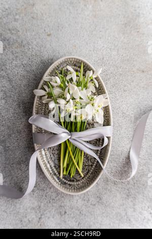 Top view of a charming display of fresh spring snowdrops artfully arranged in a vintage decorative tray, tied with a silky white ribbon, placed on a t Stock Photo