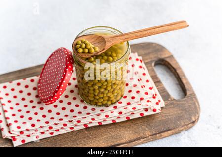 A glass jar of fresh green homemade organic peas with a wooden spoon rests on a polka dot cloth on a wooden board. The red lid with white polka dots a Stock Photo