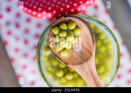 A wooden spoon holds green homemade organic peas over a glass jar, set on a white cloth with red polka dots. The vibrant colors and textures create a Stock Photo