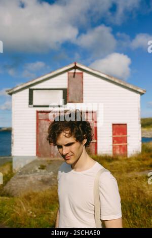 A young man with curly hair stands outdoors, smiling subtly near an old white building with red trim in Flatrock. His casual appearance and the clear, Stock Photo