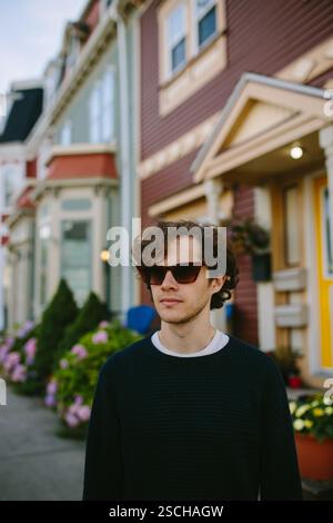 A stylish young man stands confidently on a street in St. John's, wearing sunglasses and a casual sweater. The colorful row houses in the background a Stock Photo