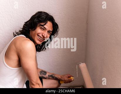 Cheerful young man with a brush in hand, painting a white wall in his home, smiling widely. His casual attire and visible tattoos add a personal touch Stock Photo