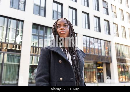 A young African American businesswoman with braids, wearing a stylish coat, takes a break outdoors in an urban setting, gazing upward thoughtfully. Stock Photo