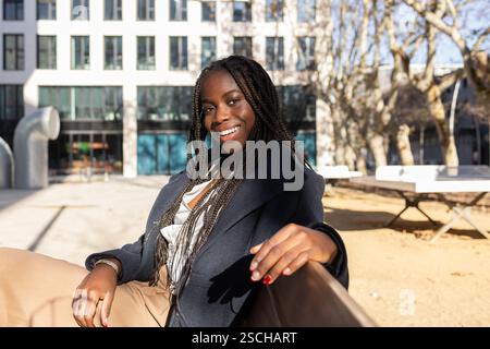 A smiling young African American businesswoman with braids relaxes outdoors during a work break sitting in a bench, reflecting positivity and relaxati Stock Photo