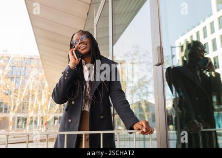 A cheerful young African American businesswoman with braids talks on her mobile phone during a break outside a modern office building. Stock Photo