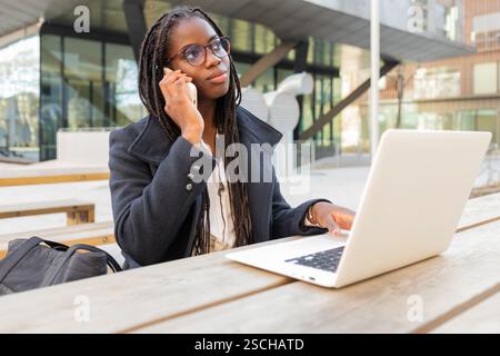 An African American businesswoman with braids is engaged in work using her laptop while taking a call on her mobile phone, seated outdoors Stock Photo