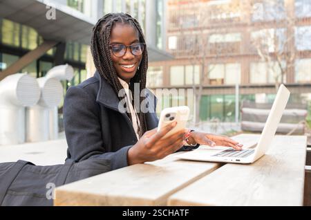 A young African American businesswoman with braids is smiling as she uses her smartphone and laptop while working remotely outdoors, capturing a momen Stock Photo