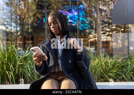 An African American businesswoman with braids intently uses a smartphone while seated outdoors on a work break, dressed in professional attire. Stock Photo