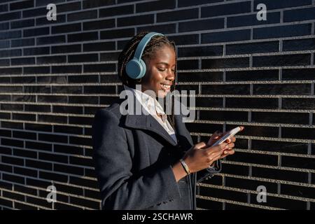 Side view of a young African American businesswoman with braids is seen enjoying a break, smiling while using her smartphone and listening to music th Stock Photo