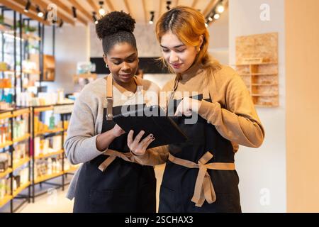 Two employees in a cosmetics store wearing aprons collaborate on a digital tablet, discussing product details. Shelves filled with diverse beauty prod Stock Photo