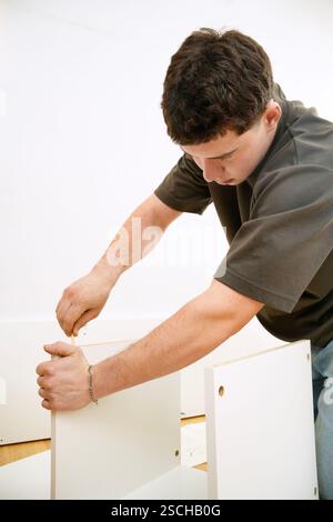 A focused young man assembles a new piece of white furniture using a screwdriver. He is seen inserting screws into the panels, showing hands-on skill Stock Photo