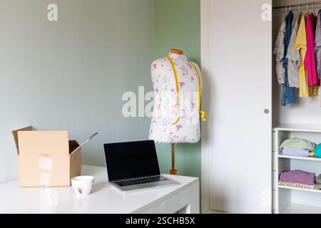 A tailor's mannequin with a floral design and measuring tape stands in a home workspace. A laptop, an open box sit on the desk, with an open closet cr Stock Photo