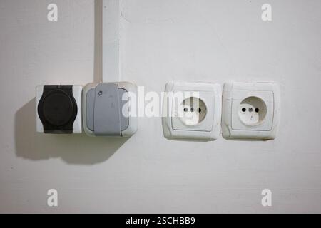 Two electrical outlets and a switch are mounted on a simple white wall. The design features a three-pin socket alongside circular two-pin sockets, sho Stock Photo