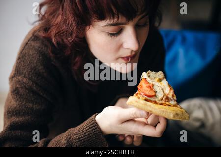 A young adult woman with curly red hair enjoys a delicious slice of pizza, focusing intently on her meal in a relaxed indoor environment. Stock Photo
