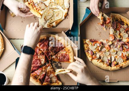 Top view of a group of unrecognizable friends gathers around the table, sharing slices of various pizzas from cardboard boxes. The image captures a ca Stock Photo