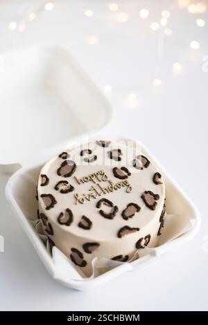 Top view of a stylish leopard print bento cake with Happy Birthday decoration in a white takeout box, surrounded by soft festive lights. Stock Photo