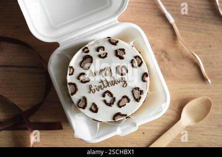 Top view of a stylish Bento birthday cake decorated with heart-shaped toppings, presented in a white takeout box with a wooden spoon alongside, set on Stock Photo