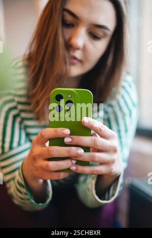 Blurred woman holding a green smartphone during a relaxing coffee break in a cozy cafe environment. Focus on hands and phone. Stock Photo