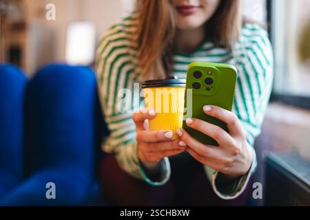 Cropped shot of an unrecognizable woman holding a smartphone and coffee cup. The focus is on her hands with vivid details in a cozy cafe setting. Stock Photo