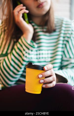Cropped image of an unrecognizable woman having a coffee break in a cafe, holding a yellow cup and speaking on a green smartphone. Stock Photo