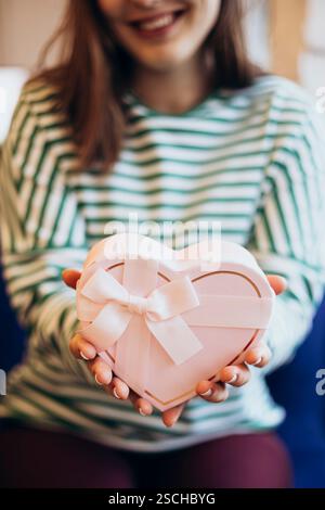 Cropped unrecognizable woman in a striped top holding a heart-shaped gift box adorned with a white ribbon, symbolizing love and generosity. Stock Photo
