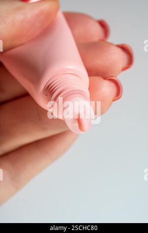 Cropped unrecognizable woman's hand applying a shiny lip balm from a pink tube, showcasing cosmetic textures on the skin in macro detail. Stock Photo