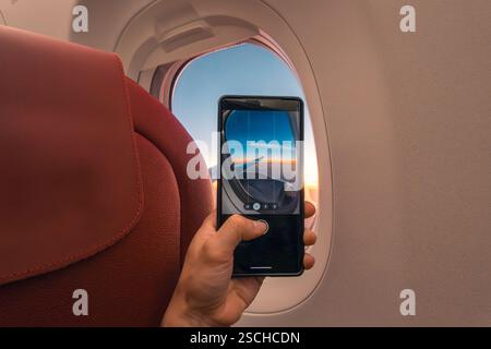 Cropped unrecognizable person captures a sunset over the ocean using a smartphone, viewed through an airplane window. Stock Photo
