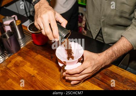 A barista pours freshly ground coffee into a paper filter placed in a pour-over coffee maker. The scene is set on a wooden counter, highlighting a spe Stock Photo