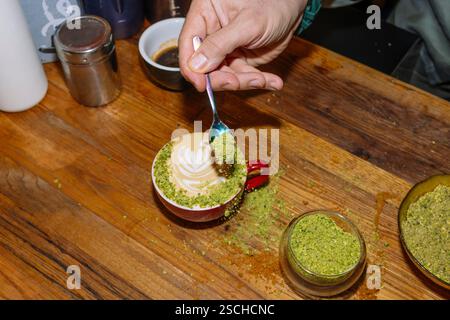 A hand sprinkles pistachio topping on a beautifully crafted latte with intricate latte art. Set on a rustic wooden table, it captures the essence of s Stock Photo