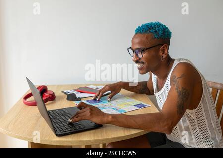 A smiling LGBTQIA+ individual with blue hair plans holidays using a laptop and map, embracing diversity and travel. The setting is casual and cheerful Stock Photo