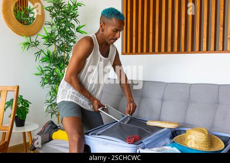 A blue-haired LGBTQIA+ man packs a suitcase on a couch for a vacation. Surrounded by travel essentials, the scene takes place in a bright, modern spac Stock Photo