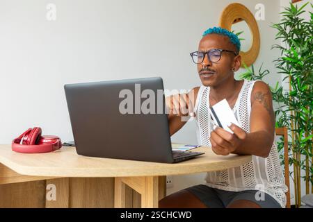 LGBTQIA+ man with blue hair using a laptop and credit card to prepare for holidays. A sense of pride and anticipation surrounds this festive moment. Stock Photo