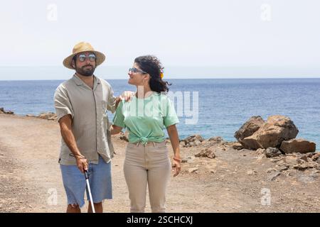 A blind man with a cane and a supportive woman walk side by side along a rocky coastal path. The serene ocean view in the background highlights their Stock Photo