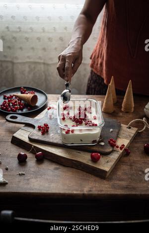 Cropped unrecognizable person scoops ice cream decorated with currants and gooseberries in a rustic kitchen setting. Stock Photo