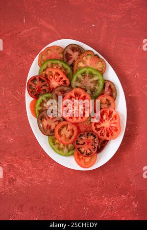 A variety of fresh heirloom tomatoes, sliced and arranged on a white plate, showcasing red, green, and brown hues. The plate sits on a textured red su Stock Photo