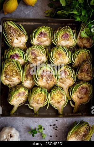 Top view of fresh artichoke halves on a baking tray, surrounded by lemons, herbs, and garlic, creating a vibrant culinary display ready for cooking. Stock Photo