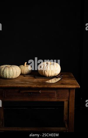 Three pumpkins rest on a rustic wooden table beside a knife, set against a dark background. The natural tones and textures create a warm, autumnal atm Stock Photo