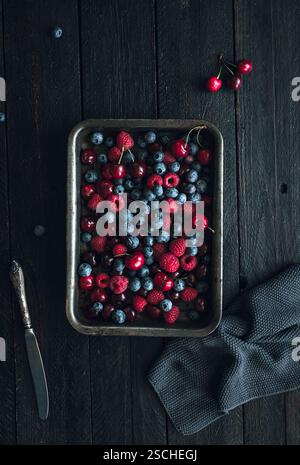A rustic tray filled with fresh blueberries, raspberries, and cherries on a dark wooden background. The vibrant colors contrast beautifully with the w Stock Photo