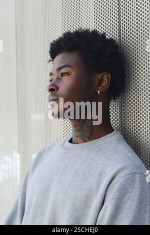 A portrait of a young black man looking away from the camera, contemplating deeply beside a textured metal wall. His expression pensive, he wears a ca Stock Photo