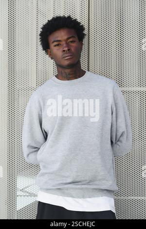 A young black man with nasal and ear piercings and distinct facial tattoos stands confidently in front of a textured wall, wearing a casual grey sweat Stock Photo