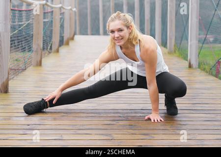 A young blond woman performs stretching exercises on a foggy wooden bridge, wearing sportswear and a focused expression. Stock Photo