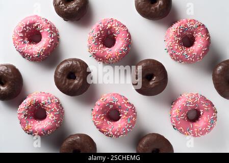 Top view of an arrangement of pink strawberry and dark chocolate donuts, each topped with colorful sprinkles, displayed on a white surface. Stock Photo