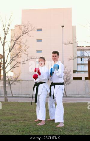 A young man and woman, both wearing karate gear with red and blue gloves respectively, practice karate outdoors with a modern building in the backgrou Stock Photo