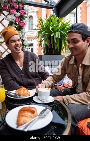 A gay couple enjoys breakfast at a cafe. They are sitting at a table with coffee and croissants, laughing and engaging in a joyful conversation. The s Stock Photo