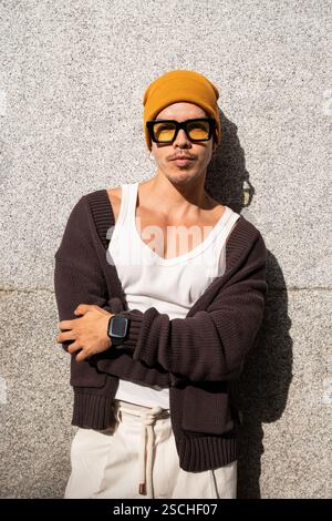 A stylish Colombian gay man leans against a concrete wall, wearing a yellow beanie, sunglasses, white tank top, and black cardigan, exuding confidence Stock Photo