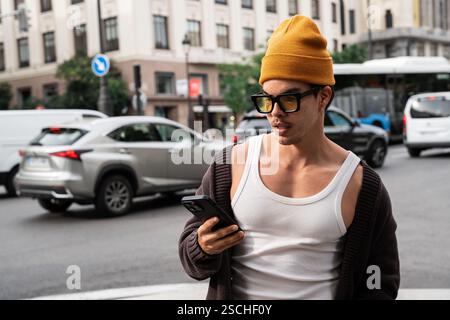 A Colombian man stands on a busy city street, wearing stylish sunglasses and a beanie as he checks his smartphone amidst bustling traffic and building Stock Photo