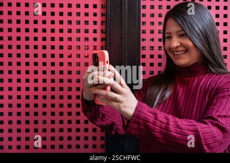 A latin woman in a maroon sweater is smiling as she takes a selfie with her smartphone against a stylish pink patterned background, capturing a joyful Stock Photo