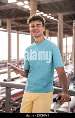 A cheerful young man stands at a train station, holding a phone and a suitcase. He is smiling and wearing a casual bright outfit, with trains visible Stock Photo