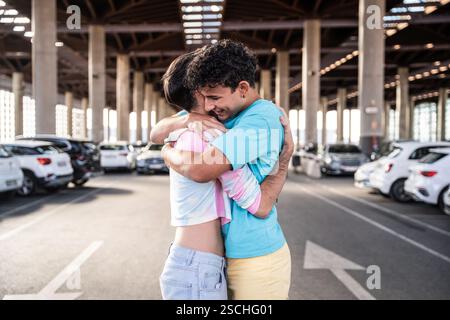 A joyful gay couple shares an embrace in a spacious urban parking garage, surrounded by parked cars. The warm moment captures love and happiness in a Stock Photo
